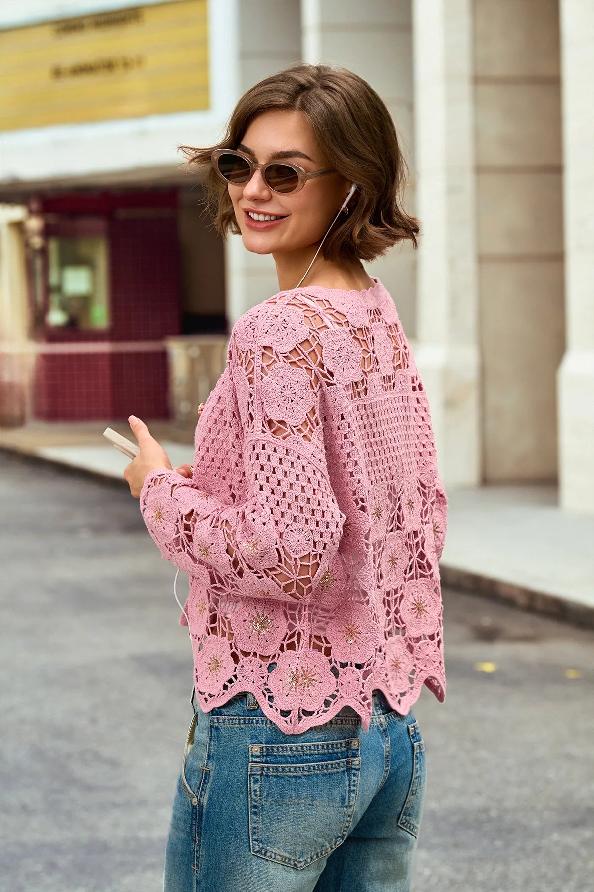 A woman wearing a pink open-knit crochet sweater and blue jeans, photographed outdoors on a city street with buildings in the background.