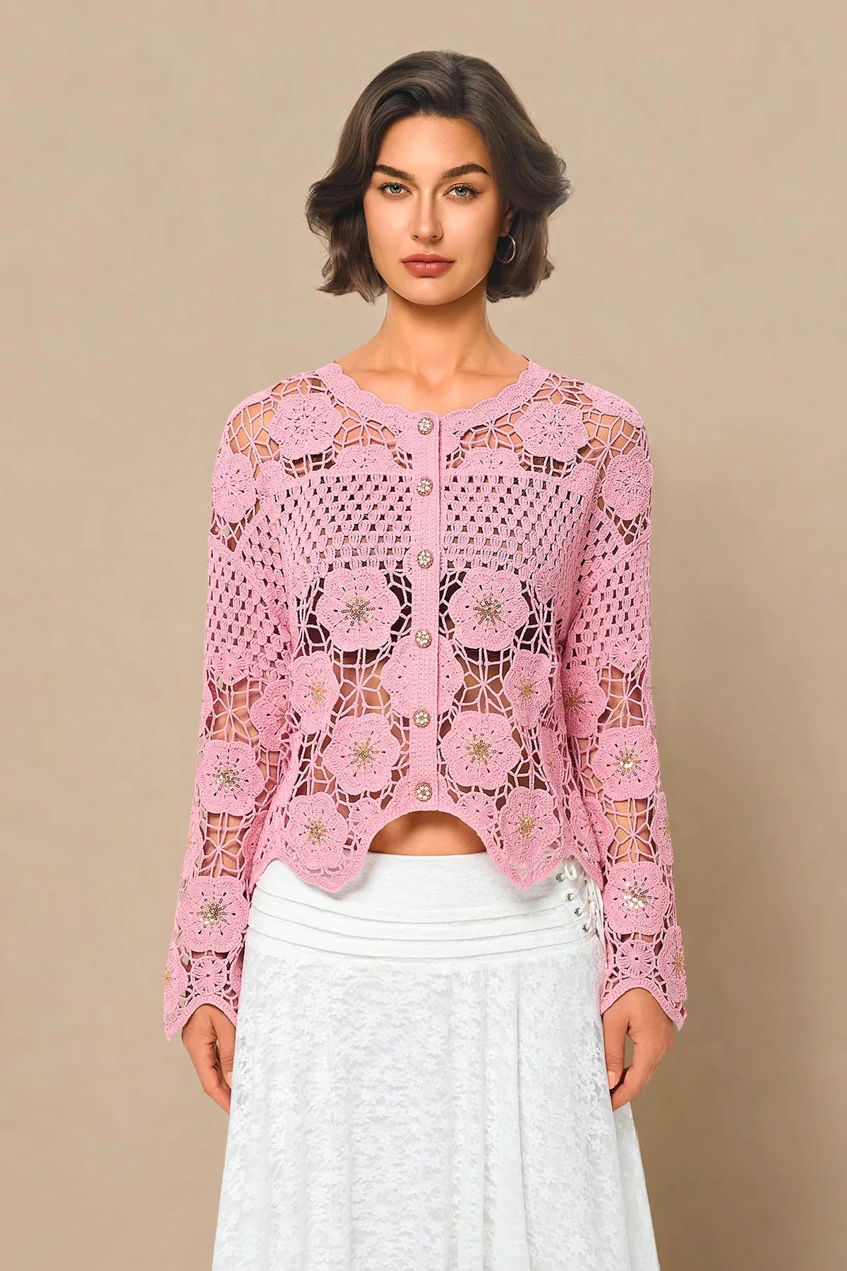 A woman modeling a pink long-sleeve crochet cardigan with a button-up front, paired with a white skirt, against a solid beige background.