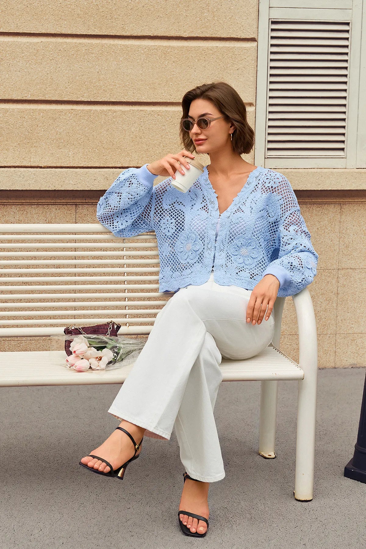 A woman sits on a white bench outdoors, wearing a light blue open-knit long-sleeve blouse with white wide-leg pants and black heeled sandals. She holds a white cup in front of her face.