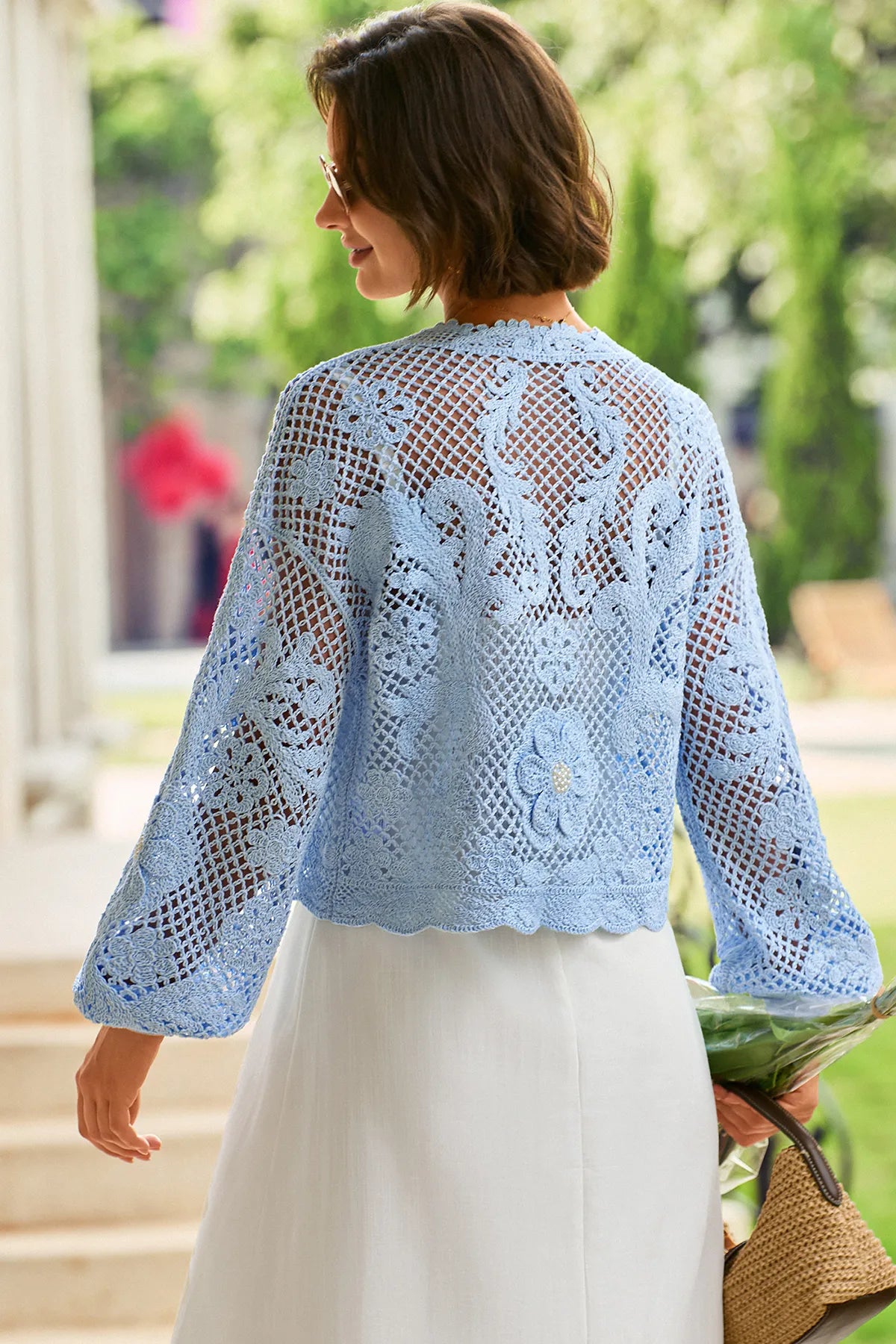 A woman shown from the back wearing a light blue crocheted lace cardigan with long sleeves and a scalloped hem, paired with a white skirt in an outdoor garden setting.