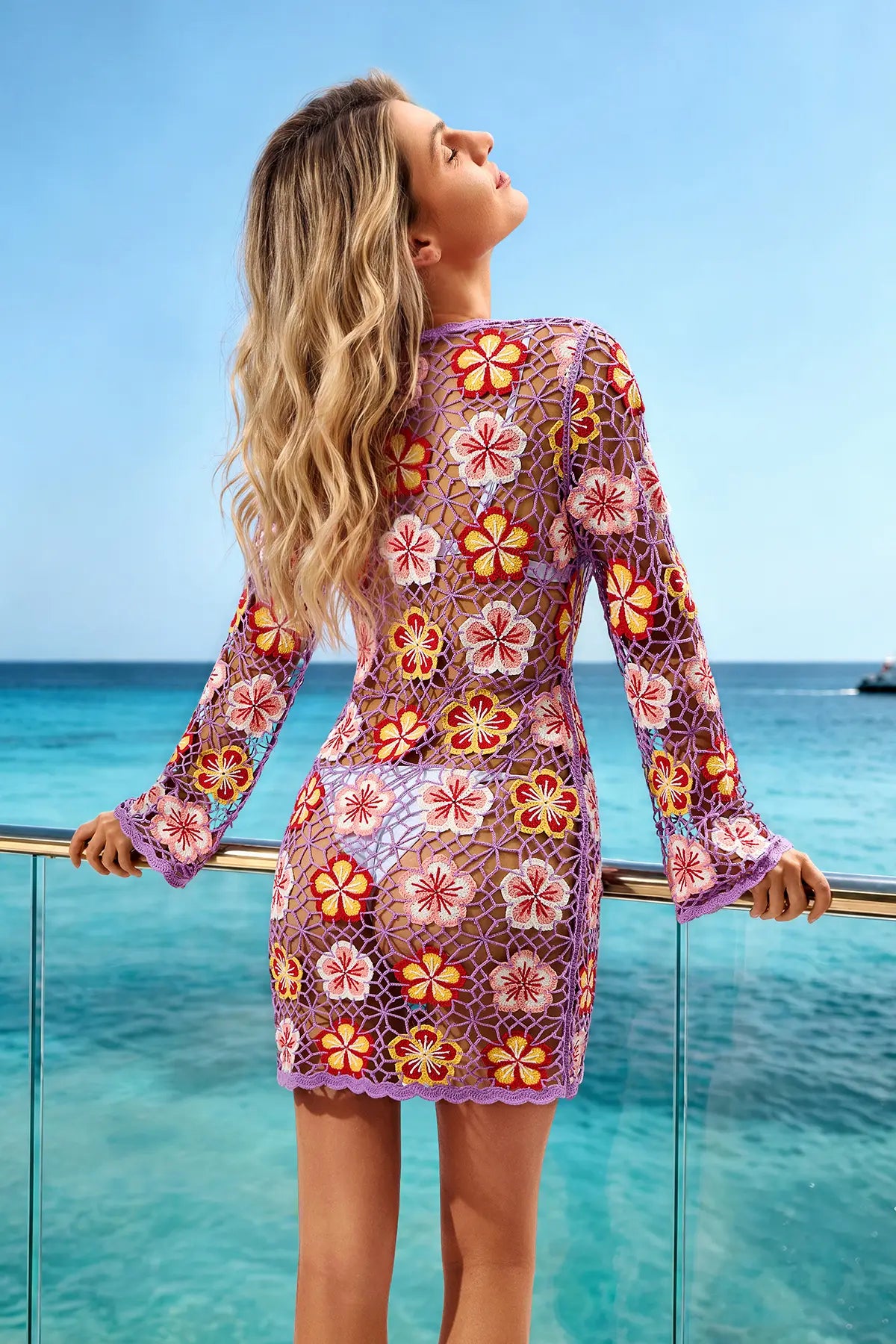 A woman wearing a long-sleeve mini dress with a colorful floral crochet lace pattern, standing on a balcony overlooking a tropical ocean under a clear blue sky.