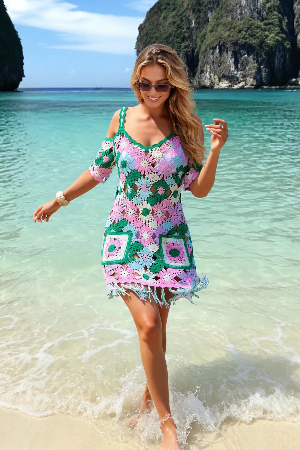 A woman wearing a colorful crochet mini dress walks into shallow turquoise ocean water on a sandy beach, with tropical cliffs in the background.