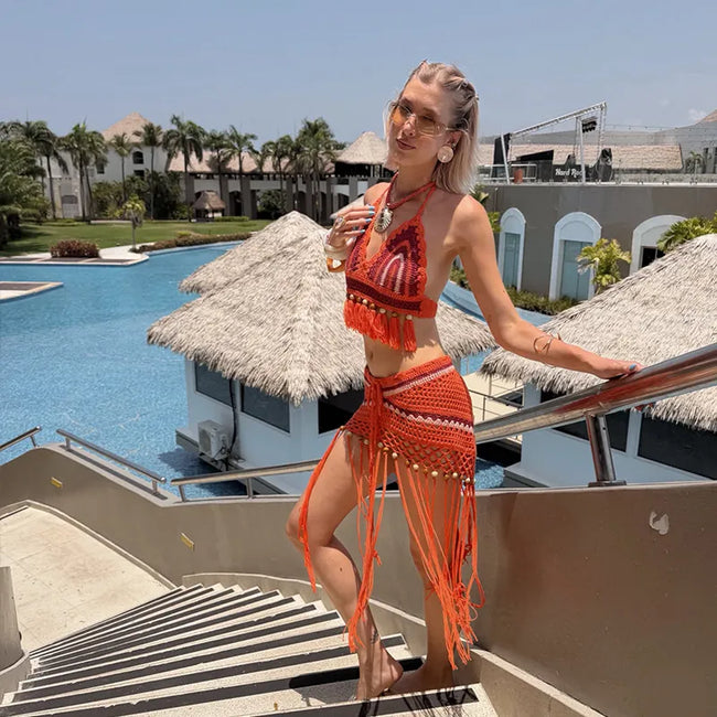 A woman wearing a bright orange crochet halter bikini top and matching fringed sarong skirt stands on outdoor resort stairs with a pool, palm trees, and thatched-roof huts in the background.