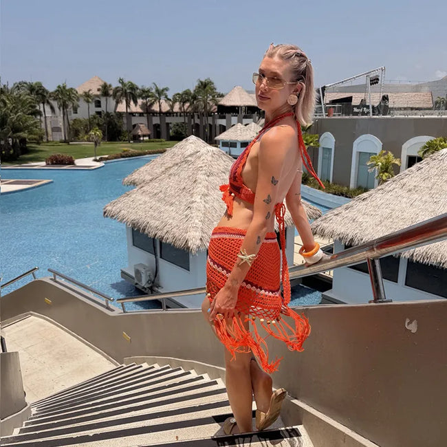 A woman wearing a bright orange crochet bikini and matching fringed wrap skirt stands on outdoor resort stairs with a swimming pool, palm trees, and thatched-roof huts in the background.