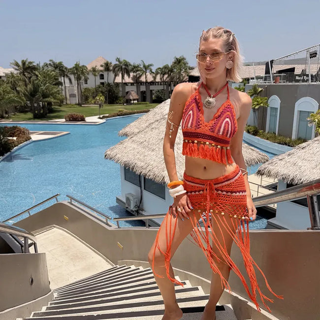 A woman wearing a bright orange and red patterned two-piece bikini with fringe details stands on outdoor resort stairs with a swimming pool, palm trees, and thatched-roof buildings in the background.