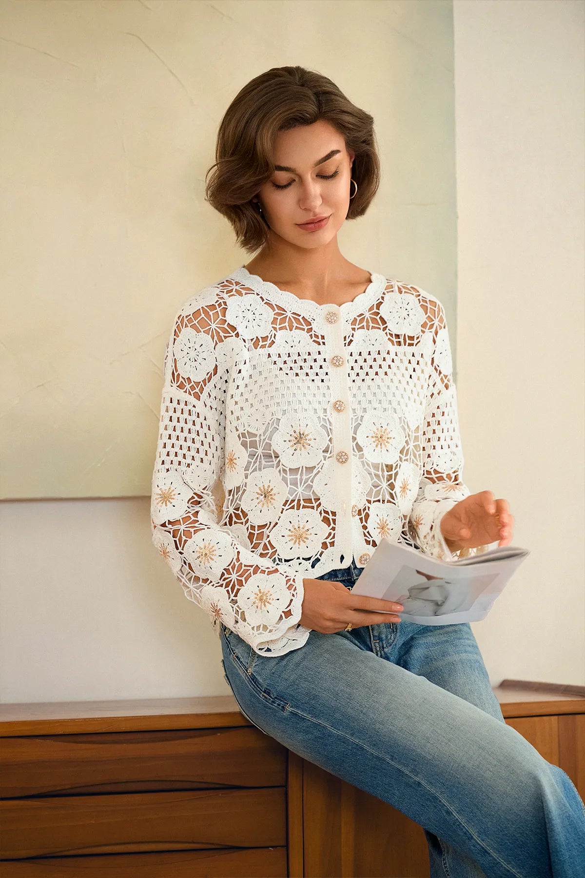 A woman with short brown hair sits on a wooden bench wearing a white long-sleeve crochet cardigan with a scalloped edge and button details, paired with blue jeans. She is holding a magazine against a neutral beige wall background.