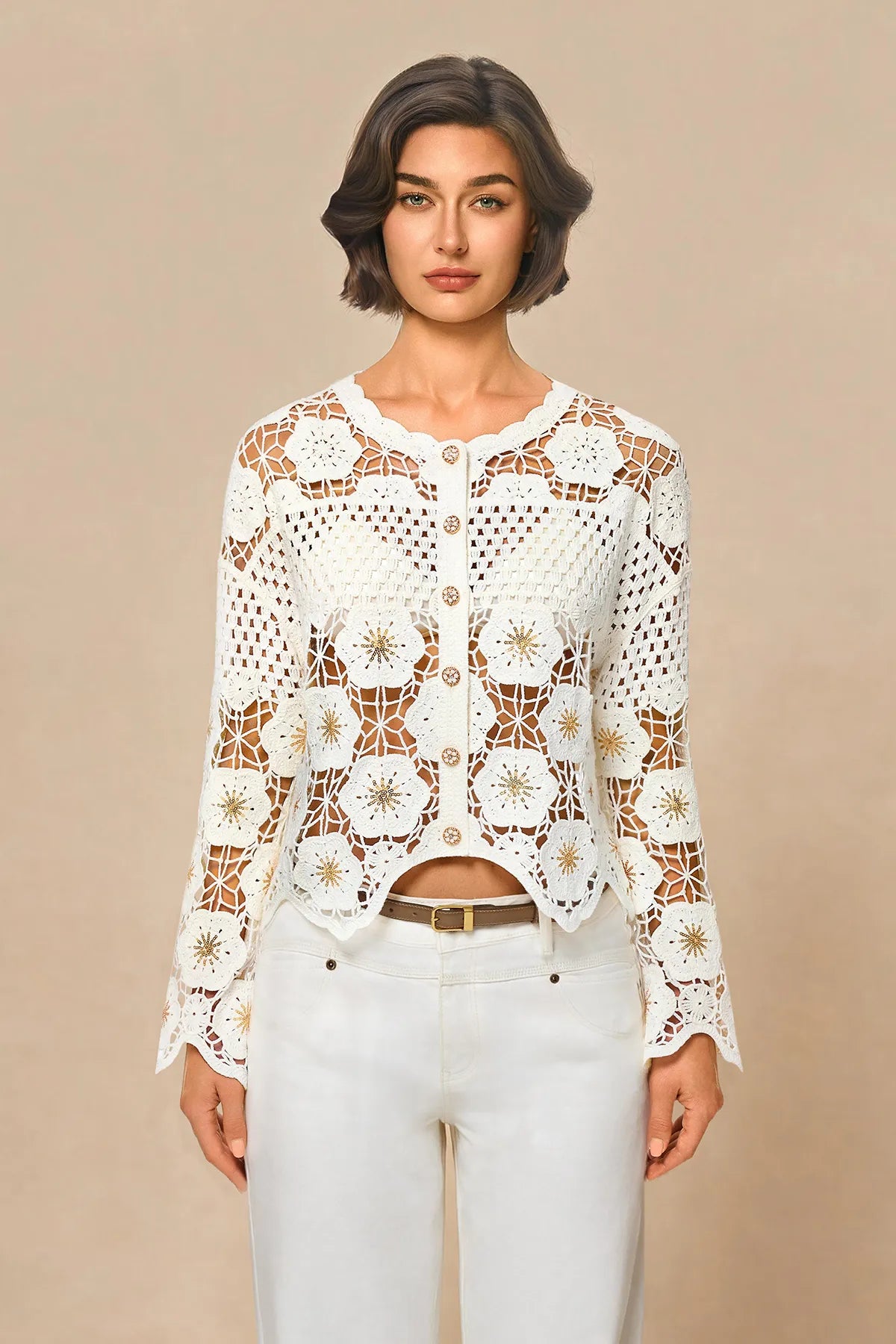A woman modeling a white long-sleeve crochet cardigan with a button-up front, paired with white pants, against a solid beige studio background.