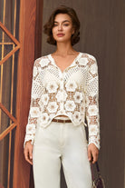 A woman models a white crochet cardigan with a V-neck and button closure, paired with white trousers and a brown belt, standing in front of a dark brown wooden door.