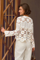 A woman wearing a white floral crochet lace long-sleeve top and cream trousers, standing in front of a wooden door and holding a small brown handbag.