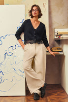 A woman modeling a black long-sleeve cardigan with a lace hem and beige wide-leg trousers in a styled indoor room with abstract wall art and a wooden floor.