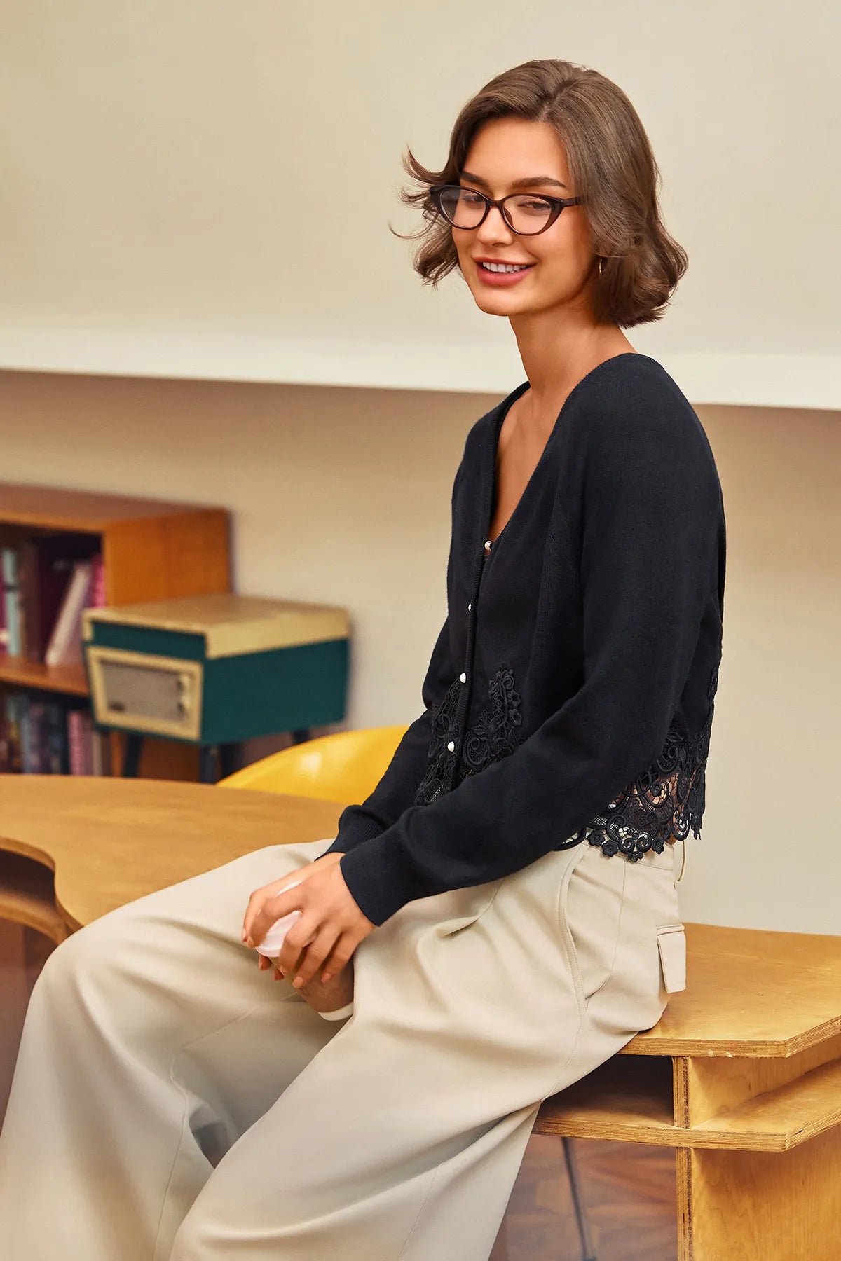 A woman with short brown hair and glasses sits on a wooden bench in a warm indoor setting, wearing a black lace-trim cardigan and light beige wide-leg trousers.