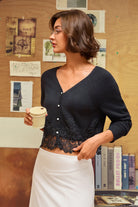 A woman wearing a black lace-trimmed cardigan and a white skirt, holding a paper coffee cup in front of a wooden bulletin board.