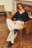 A woman wearing a black long-sleeve knit cardigan with a lace hem, cream wide-leg trousers, and black loafers sits on a yellow chair in a warm, wood-toned room.