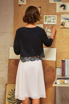 A woman viewed from behind wearing a black cropped cardigan with a lace hem and a white skirt, standing in front of a wooden board with pinned artwork in an indoor setting.