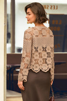 A woman wearing a beige crochet lace long-sleeve top and a brown skirt, photographed from the back in an indoor setting with a blurred background.