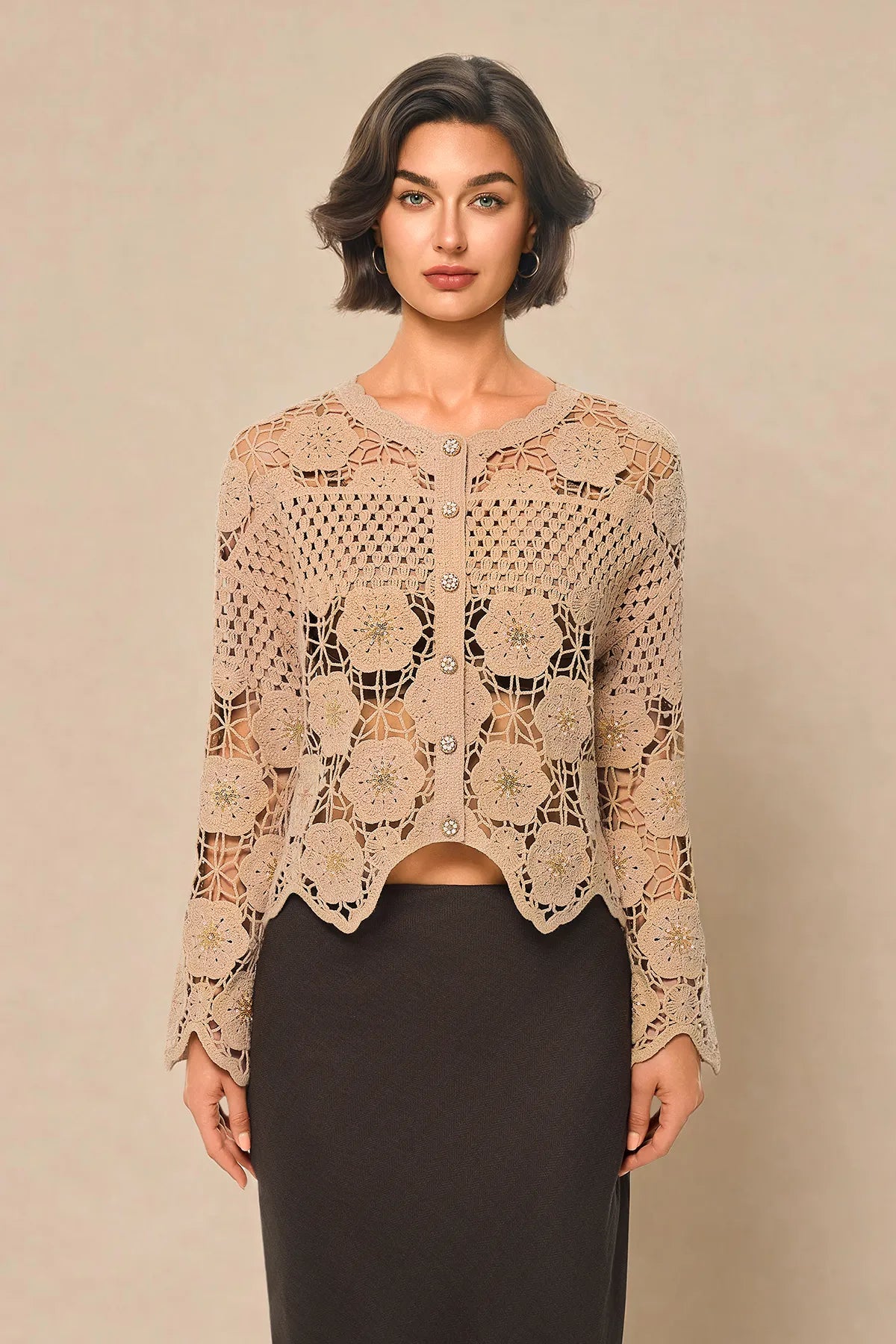 A woman modeling a beige crochet cardigan with long sleeves and a button front, paired with a dark skirt, against a solid beige studio background.