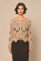 A woman modeling a beige crochet cardigan with long sleeves and a button front, paired with a dark skirt, against a solid beige studio background.