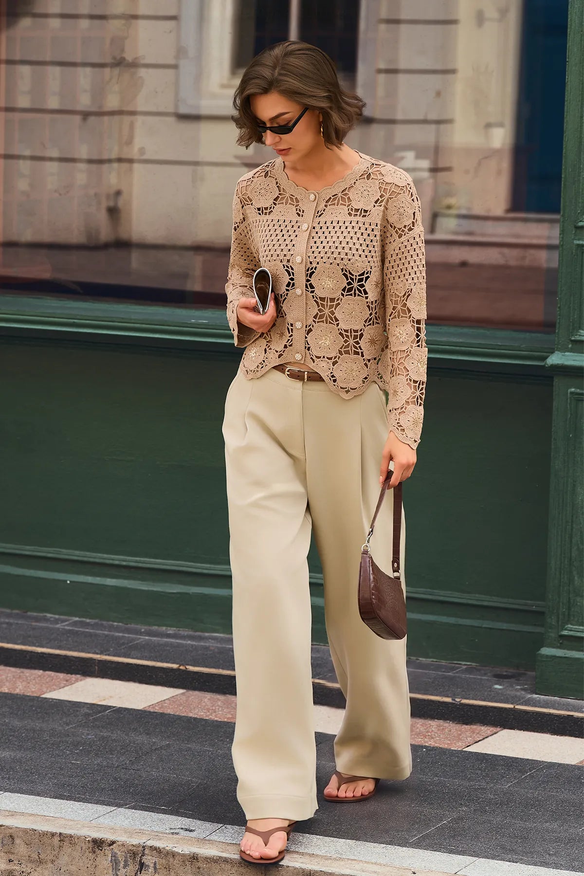 A woman walking on a city sidewalk wearing a beige crochet cardigan, wide-leg beige trousers, and carrying a brown handbag.