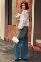 A woman wearing a white crochet long-sleeve top and blue wide-leg jeans, holding a small white handbag and pink flowers, standing outdoors against a brick wall.