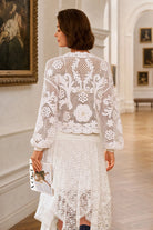 A woman wearing a white crochet lace long-sleeve cropped jacket and matching white lace skirt, photographed from the back in an elegant art gallery hallway.
