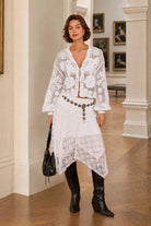 A woman models a white crochet knit three-piece set consisting of a cropped long-sleeve top, a matching asymmetrical skirt, and a black buttoned belt, styled with black leather ankle boots and a black handbag in an indoor hallway.