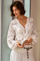 A woman wearing a white crochet cardigan and matching white skirt, holding an open book. The cardigan features intricate floral lace crochet patterns and a V-neckline.