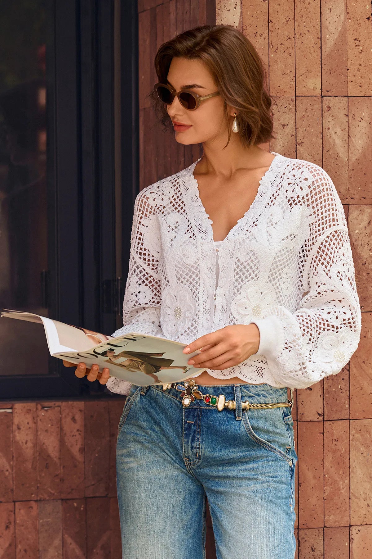 A woman wearing a white long-sleeve crochet lace blouse and blue jeans stands against a brown tiled wall, holding an open magazine.