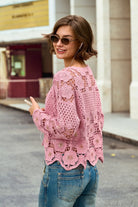 A woman wearing a pink open-knit crochet sweater and blue jeans, photographed outdoors on a city street with buildings in the background.