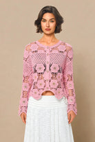 A woman modeling a pink long-sleeve crochet cardigan with a button-up front, paired with a white skirt, against a solid beige background.