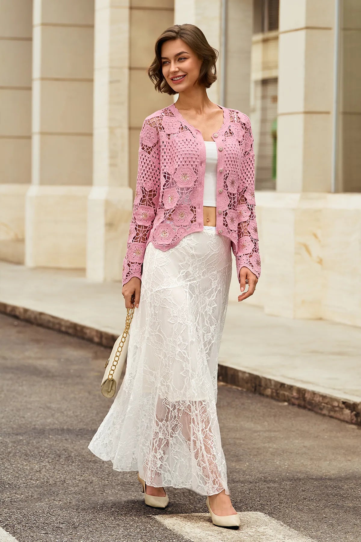 A woman stands outdoors in front of a stone building wearing a pink crochet cardigan over a white top and a long white lace skirt, holding a beige handbag.