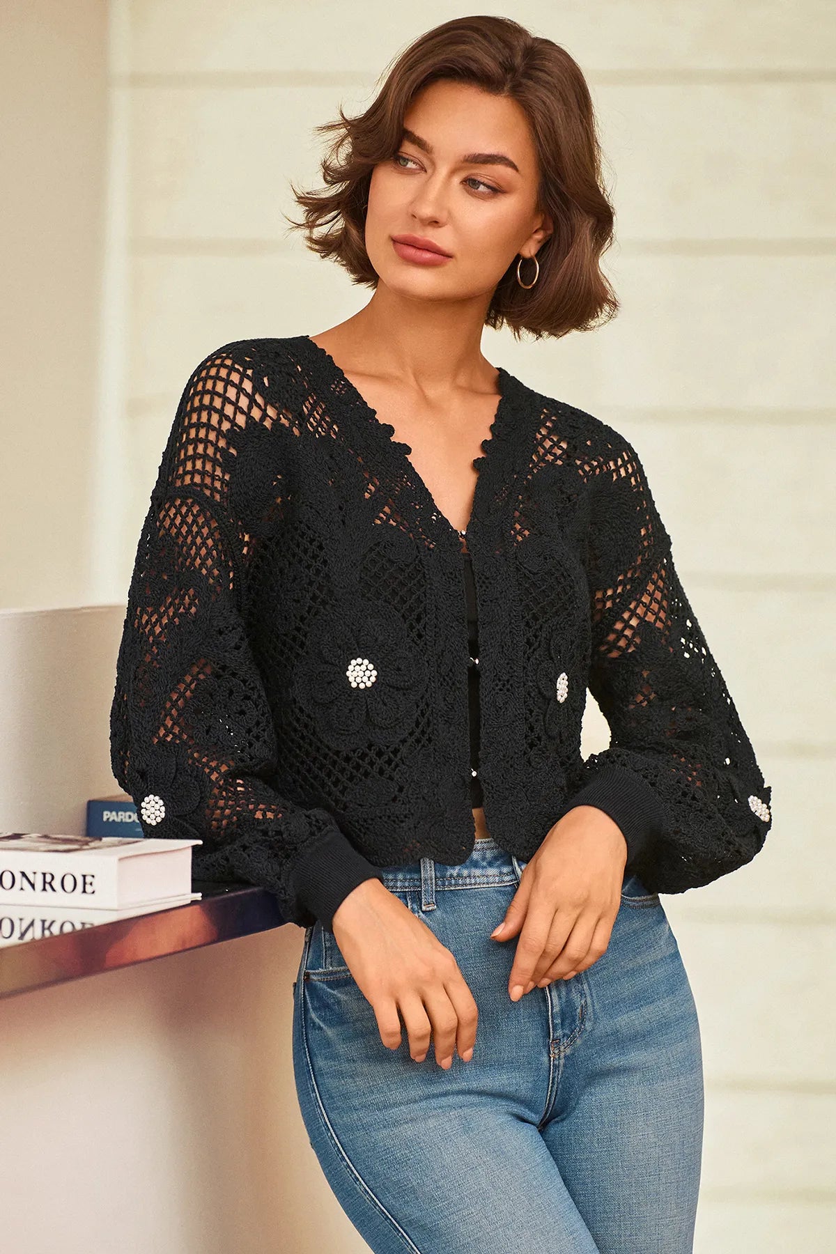 A woman wearing a black open-knit cardigan with decorative silver floral embellishments, paired with blue jeans, leaning against a light-colored wall next to a book.