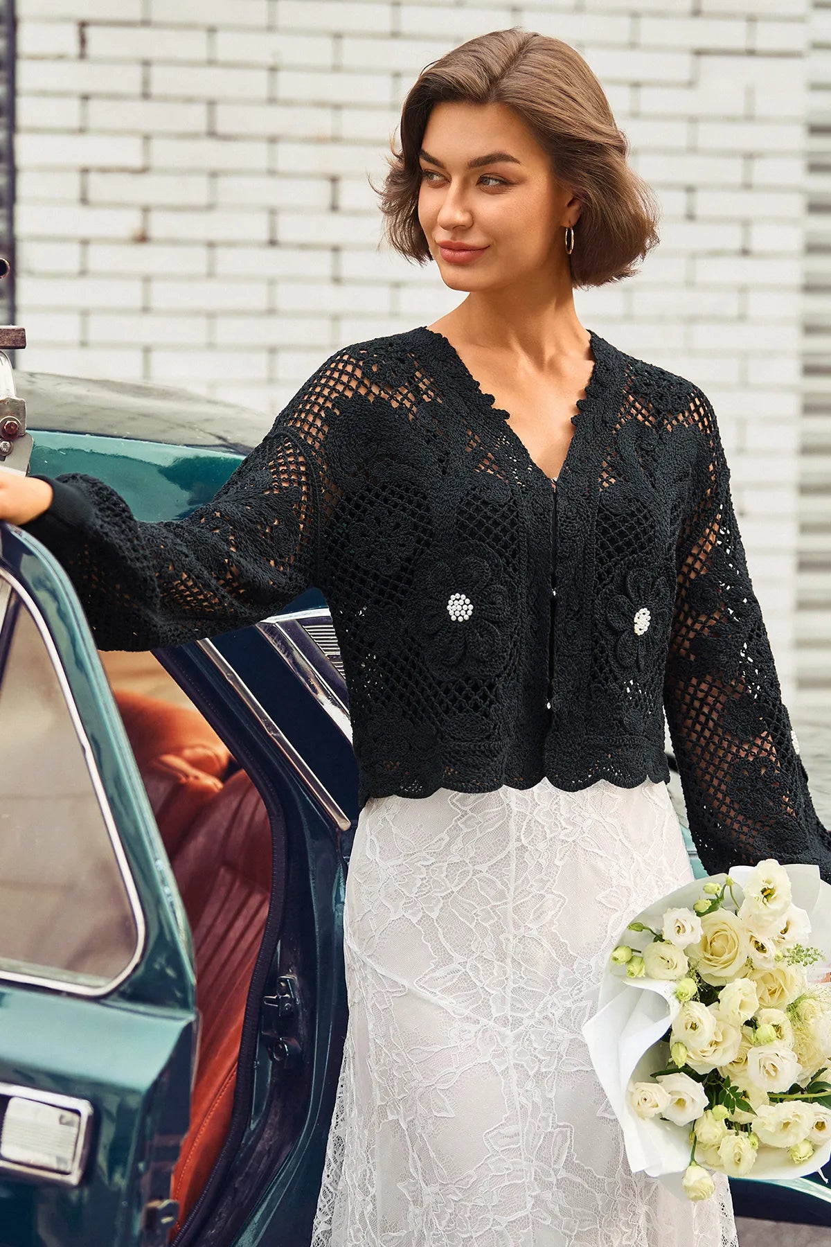 A woman stands beside a vintage teal car wearing a black crocheted cardigan with floral appliqués and a white lace dress, holding a bouquet of white roses against a white brick wall.