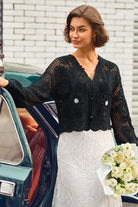 A woman stands beside a vintage teal car wearing a black crocheted cardigan with floral appliqués and a white lace dress, holding a bouquet of white roses against a white brick wall.