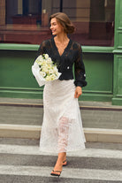 A woman walking outdoors in a black lace long-sleeve top and a white lace midi skirt, holding a bouquet of white flowers.