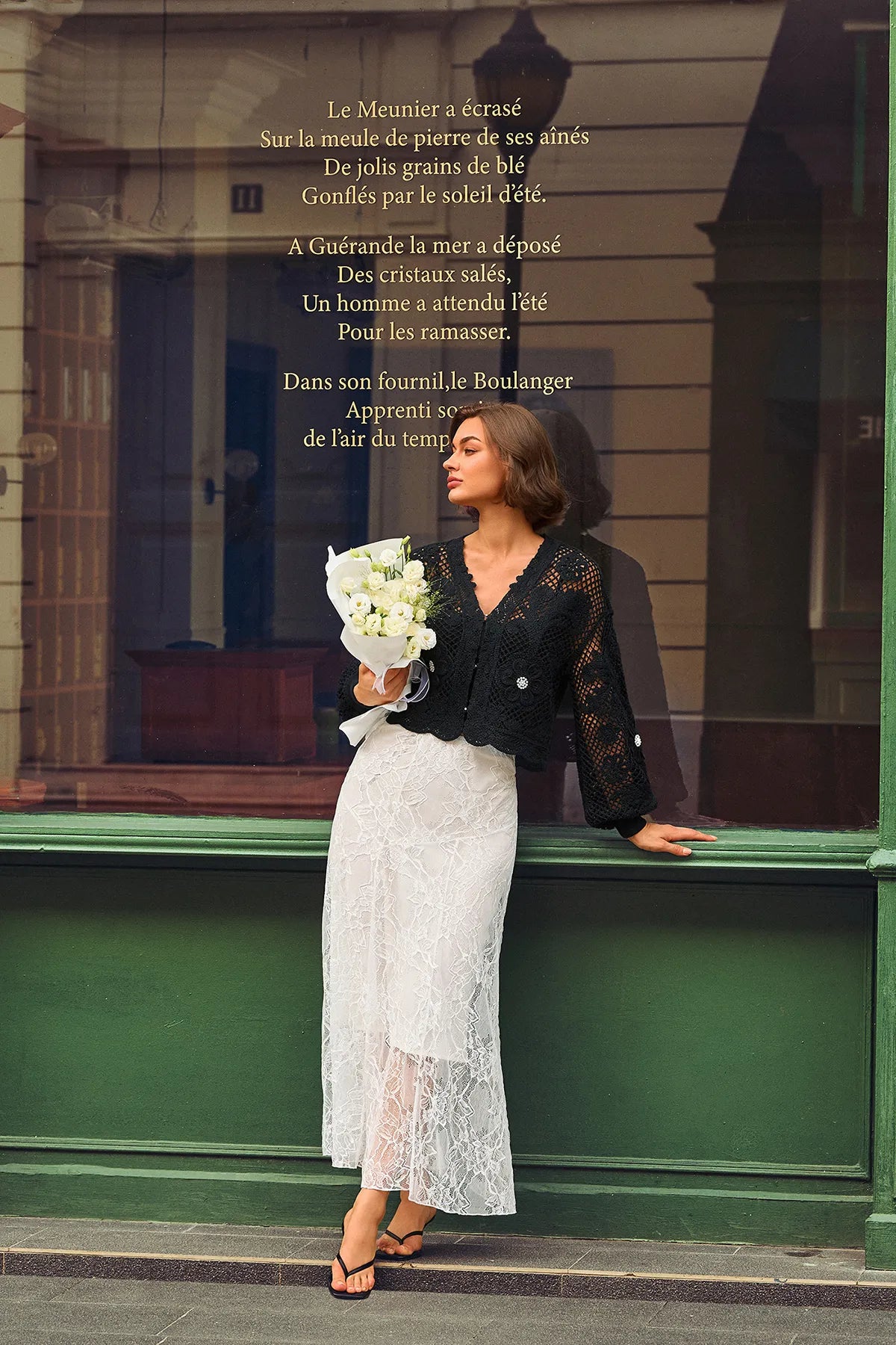 A woman leans against a green storefront window holding a bouquet of white flowers. She is wearing a black lace long-sleeve top and a white lace midi skirt.