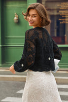 A woman wearing a black lace cropped cardigan with silver embellishments and a white lace skirt, photographed outdoors in front of a green building.
