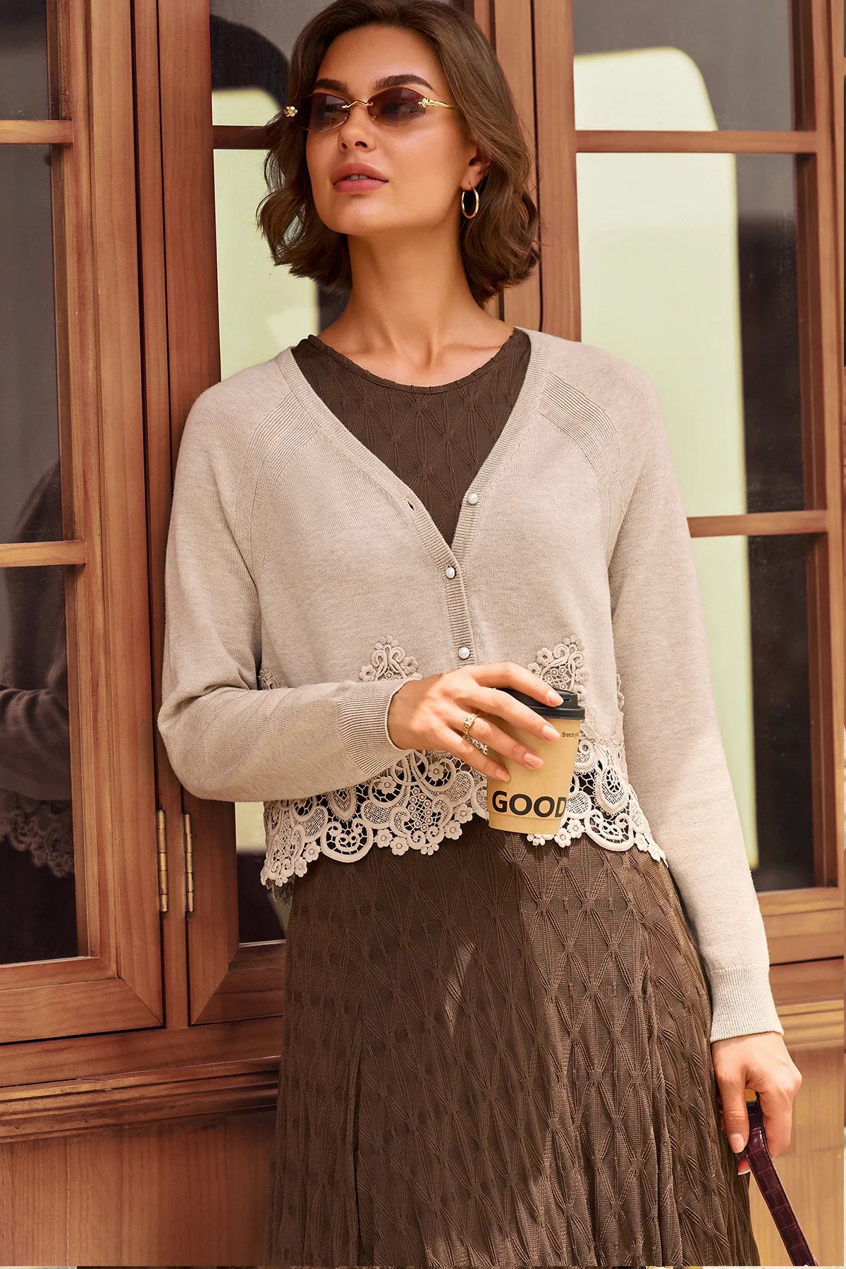 A woman models a light beige cardigan with a brown lace hem detail, layered over a brown knit top and a brown pleated skirt, standing in front of a wooden window frame.