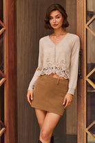 A woman models a light beige knit cardigan with a delicate lace hem detail, paired with a brown mini skirt, standing in front of a rustic wooden door.