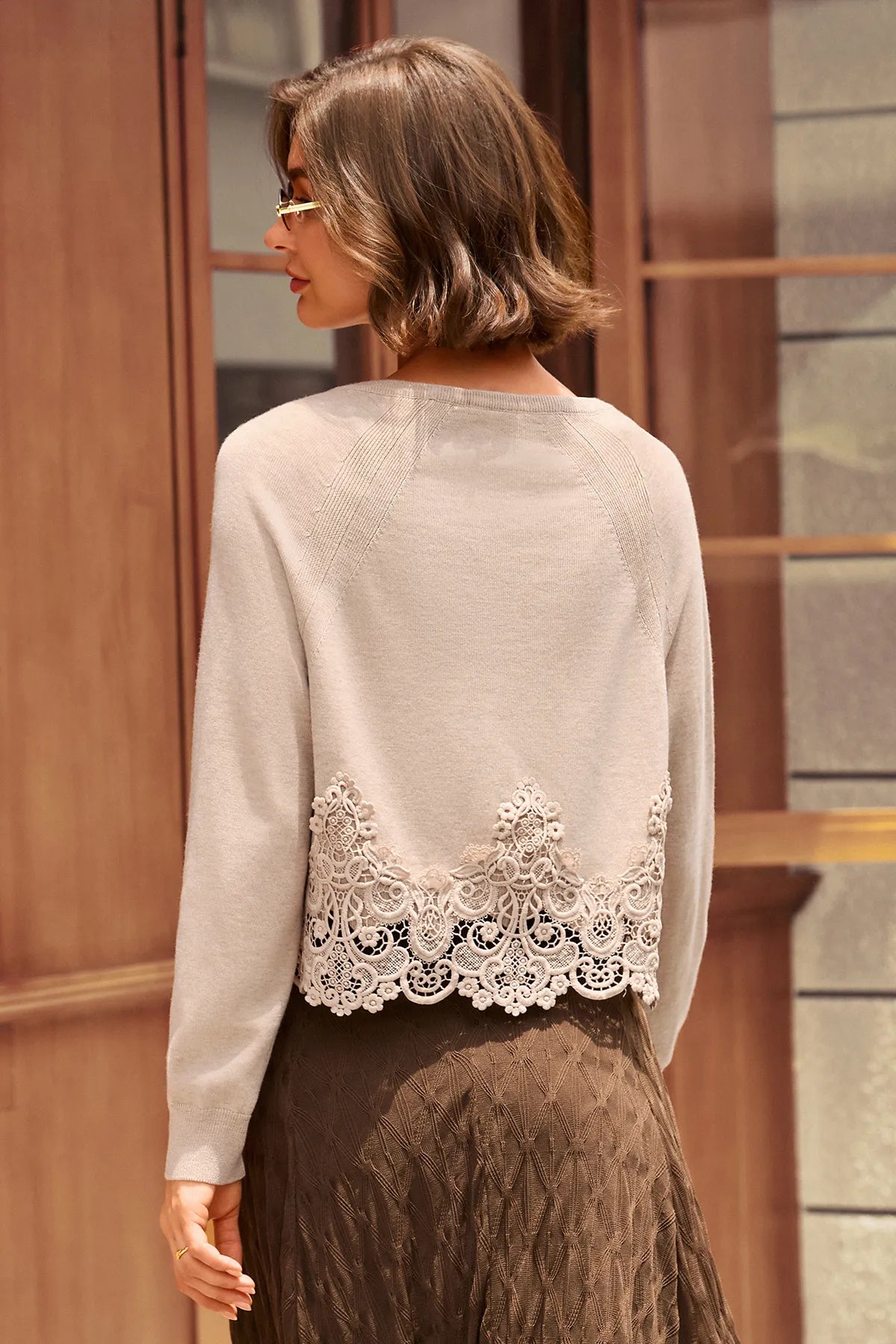 A woman shown from the back wearing a light beige knit cardigan with a prominent white lace hem, paired with a brown skirt, set against a warm-toned wooden building background.