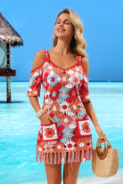 A woman stands on a tropical beach wearing a red crochet cold-shoulder mini dress with white and light blue floral crochet patterns and fringe trim. She is holding a woven straw tote bag.