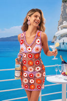A woman stands on a yacht balcony wearing a sleeveless crochet mini dress with a floral pattern in orange, red, and lavender. She holds a bottle and a cocktail glass, with a table of drinks and a blue ocean background.