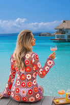 Woman in a floral dress holding a glass of pink wine by a turquoise ocean with a resort in the background.