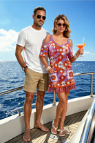 A man and a woman standing on a yacht deck with the ocean and blue sky behind them. The woman is wearing a colorful floral crochet-style mini dress with fringe trim and holding an orange cocktail.