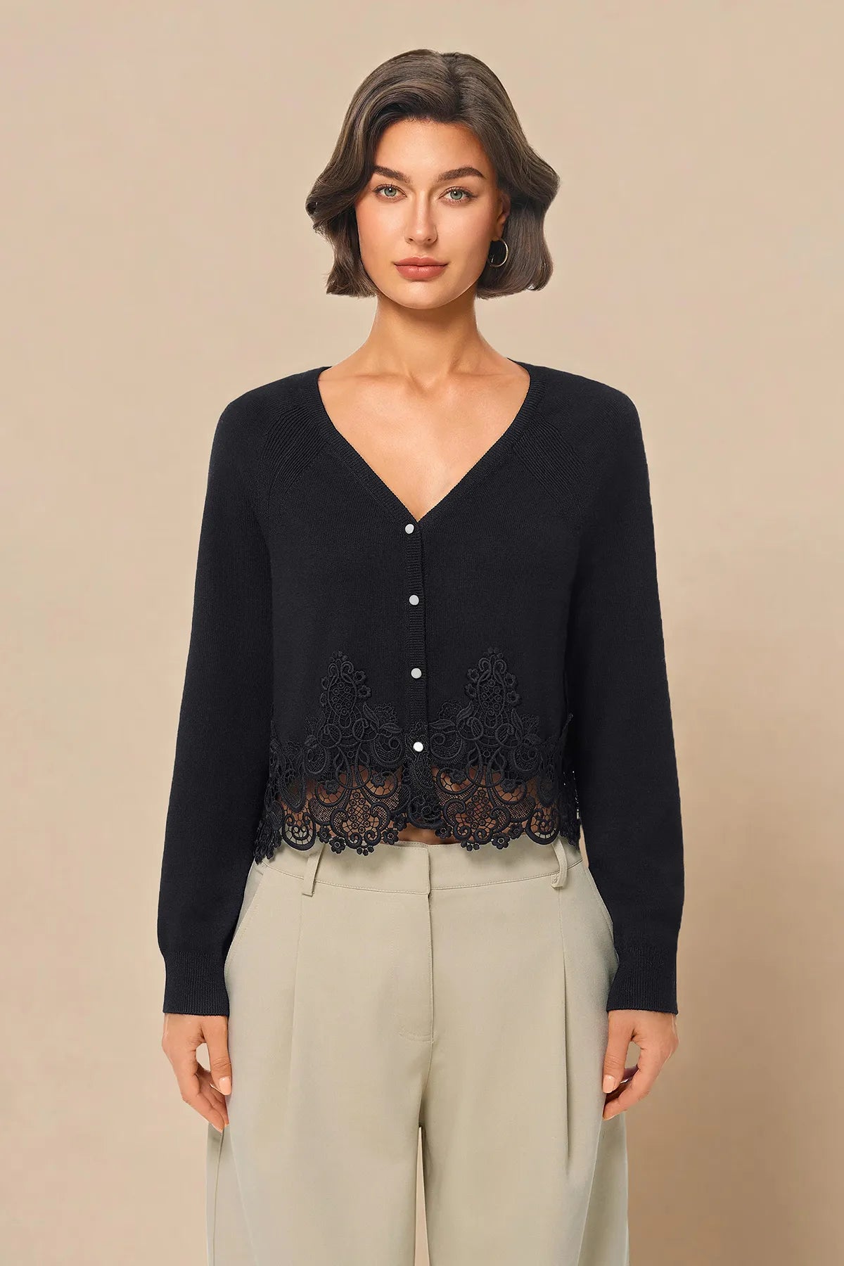 A woman modeling a black long-sleeve V-neck cardigan with a lace hem detail and light beige wide-leg trousers, set against a solid beige studio background.