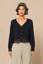 A woman modeling a black long-sleeve V-neck cardigan with a lace hem detail and light beige wide-leg trousers, set against a solid beige studio background.