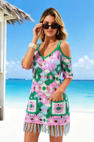 A woman wearing a colorful crochet cold-shoulder mini dress with fringe, standing on a sunny beach with turquoise water and a white sand background.