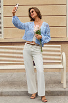 A woman takes a selfie outdoors in front of a beige building and bench, wearing a light blue crochet cropped cardigan over a white top, white wide-leg jeans, and black sandals, while holding pink flowers.