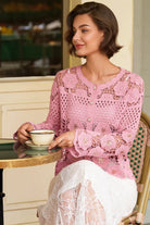 A woman seated at a cafe table wearing a pink crocheted cardigan with floral lace detailing and a white lace skirt, holding a teacup.