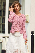 A woman wearing a pink long-sleeve crochet cardigan with floral lace patterns over a white skirt, standing in front of a white building wall.