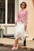 A woman models a pink crochet long-sleeve crop top and a white lace midi skirt, standing outdoors on a sidewalk next to a building with a window and bookshelves.